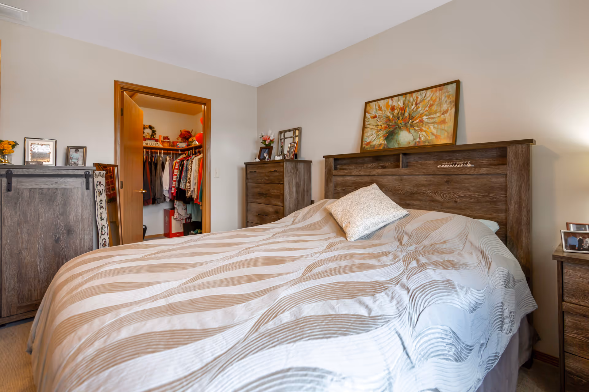 A furnished bedroom with a large bed, wooden headboard and dressers, and an open closet visible through a doorway.