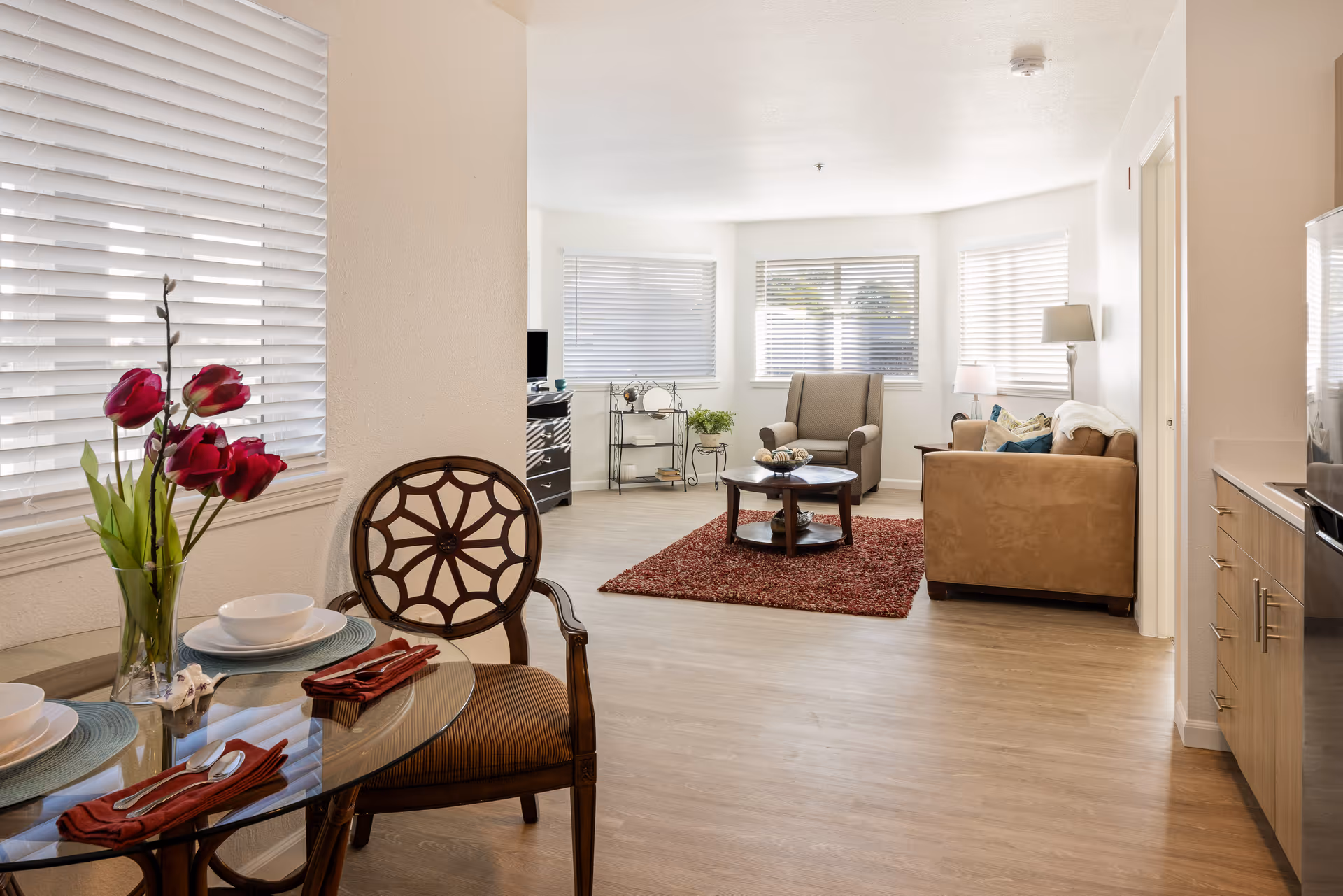 A bright and cozy senior living community interior featuring a small dining area with a glass table set for two, decorated with a vase of red flowers. The space opens into a living room with a beige sofa, an armchair, a round coffee table on a red rug, and large windows with white blinds allowing natural light to fill the room.