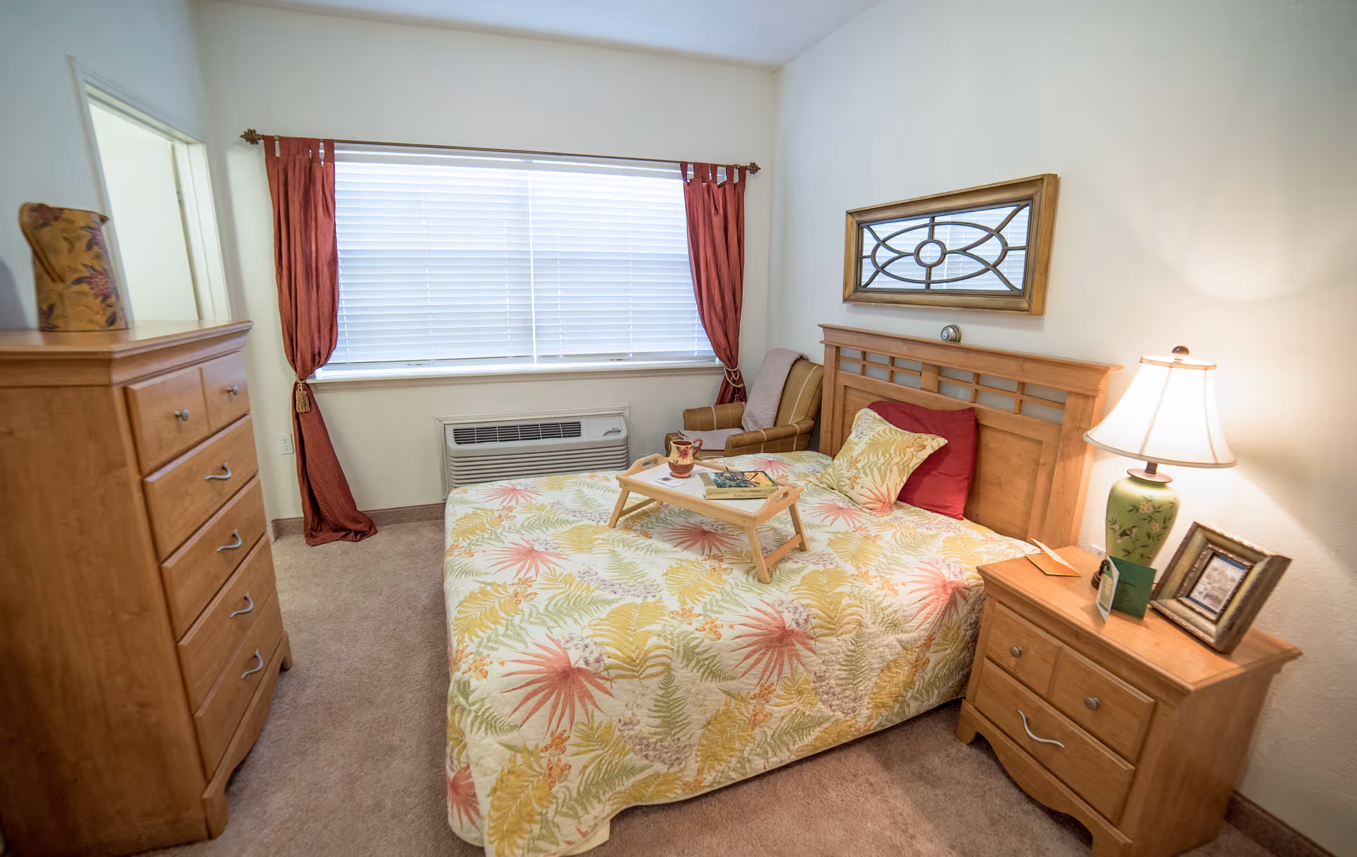 Sunlit furnished bedroom with a bed covered in a floral quilt, wooden headboard and nightstand with lamp, dresser, window with blinds, and an armchair.