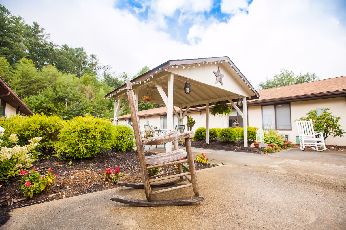 Covered entrance of a care facility with a rustic wooden rocking chair in the foreground, landscaped shrubs, and a porch area with seating.