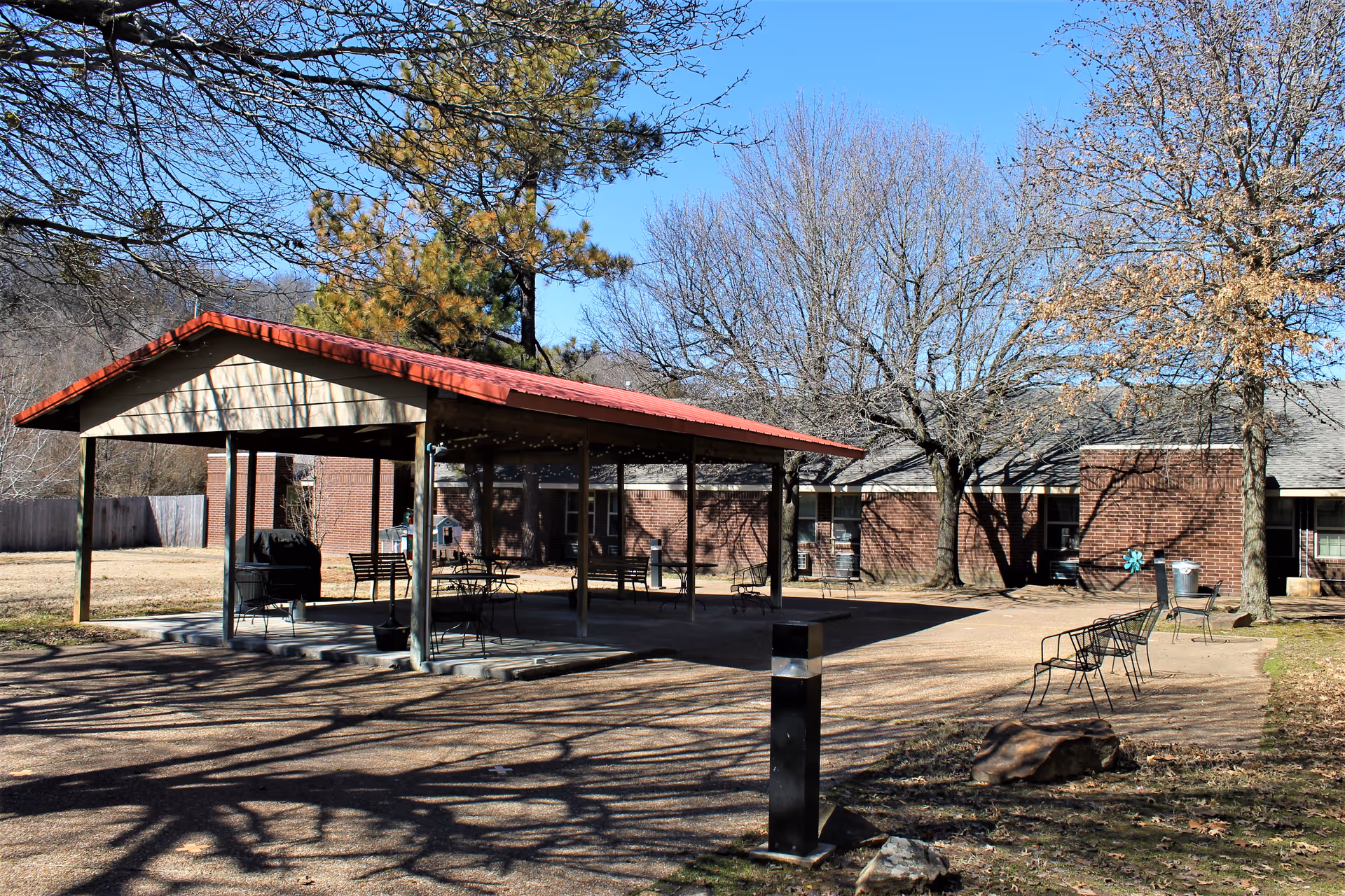 Outdoor courtyard with a covered pavilion, tables and chairs in front of a brick nursing home building and leafless trees.
