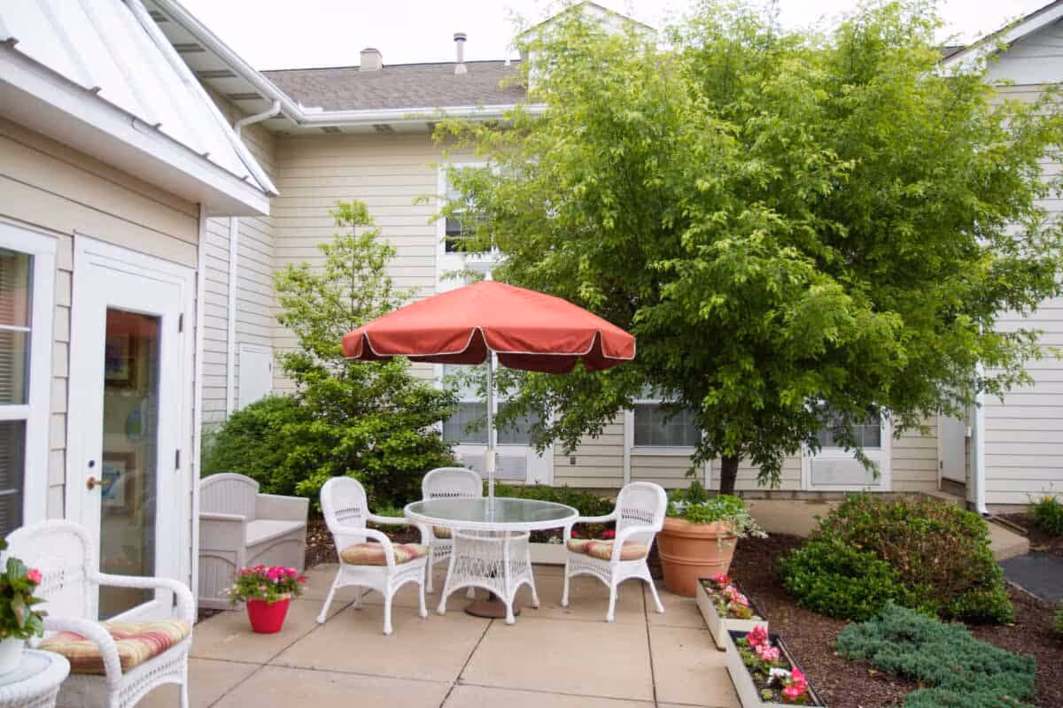 Outdoor patio area with white wicker chairs and a glass-top table under a red umbrella. The patio is surrounded by green bushes, potted plants, and trees, adjacent to a beige building with windows and a door.