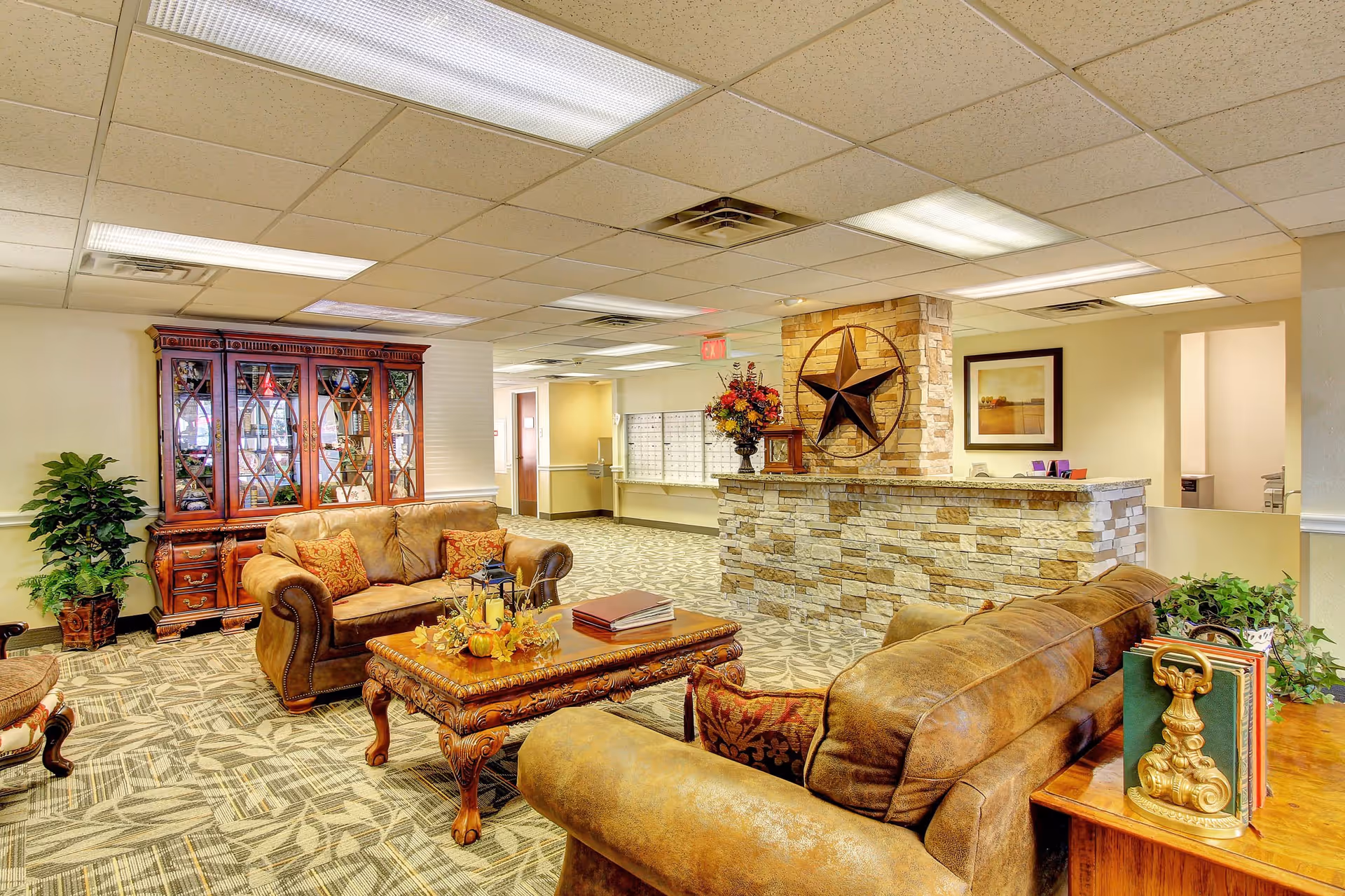 A cozy and well-lit common area in a senior living facility featuring two brown leather sofas with decorative pillows, an ornate wooden coffee table with fall-themed decorations, a wooden cabinet with glass doors, a stone reception desk with a large star decoration on the wall behind it, and a patterned carpet. There are plants and framed artwork adding to the warm ambiance.