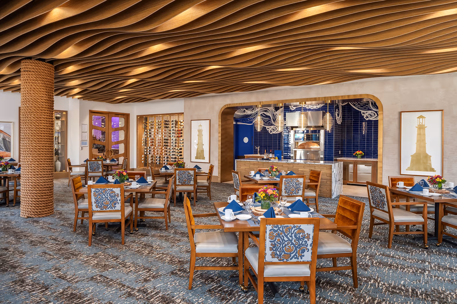 A spacious dining room with wooden tables and chairs, each table set with blue napkins, white cups, plates, and glassware. The ceiling features a unique wave-like wooden design with recessed lighting. A pillar wrapped in thick rope stands on the left side. The back wall has a wine rack and a kitchen area with blue tiled walls and pendant lights. Framed artwork of lighthouses is displayed on the walls.