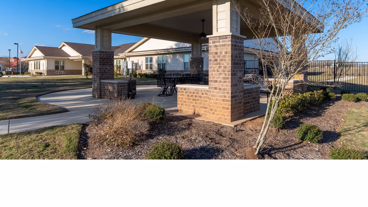 Outdoor covered seating area with brick pillars and metal tables and chairs, surrounded by landscaped bushes and trees, with single-story residential buildings in the background under a clear blue sky.