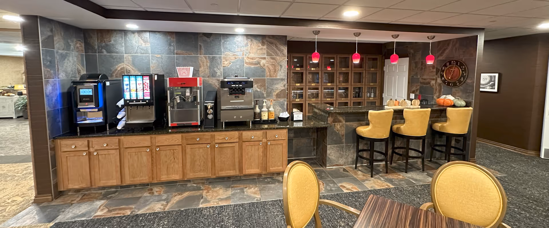 A beverage and snack station area with a coffee machine, soda dispenser, popcorn machine, and condiment bottles on a granite countertop with wooden cabinets below. To the right, there is a bar counter with three yellow cushioned bar stools and three red pendant lights hanging above. The wall behind the bar has a clock and wooden cubby shelves. In the foreground, part of a table and yellow cushioned chairs are visible.