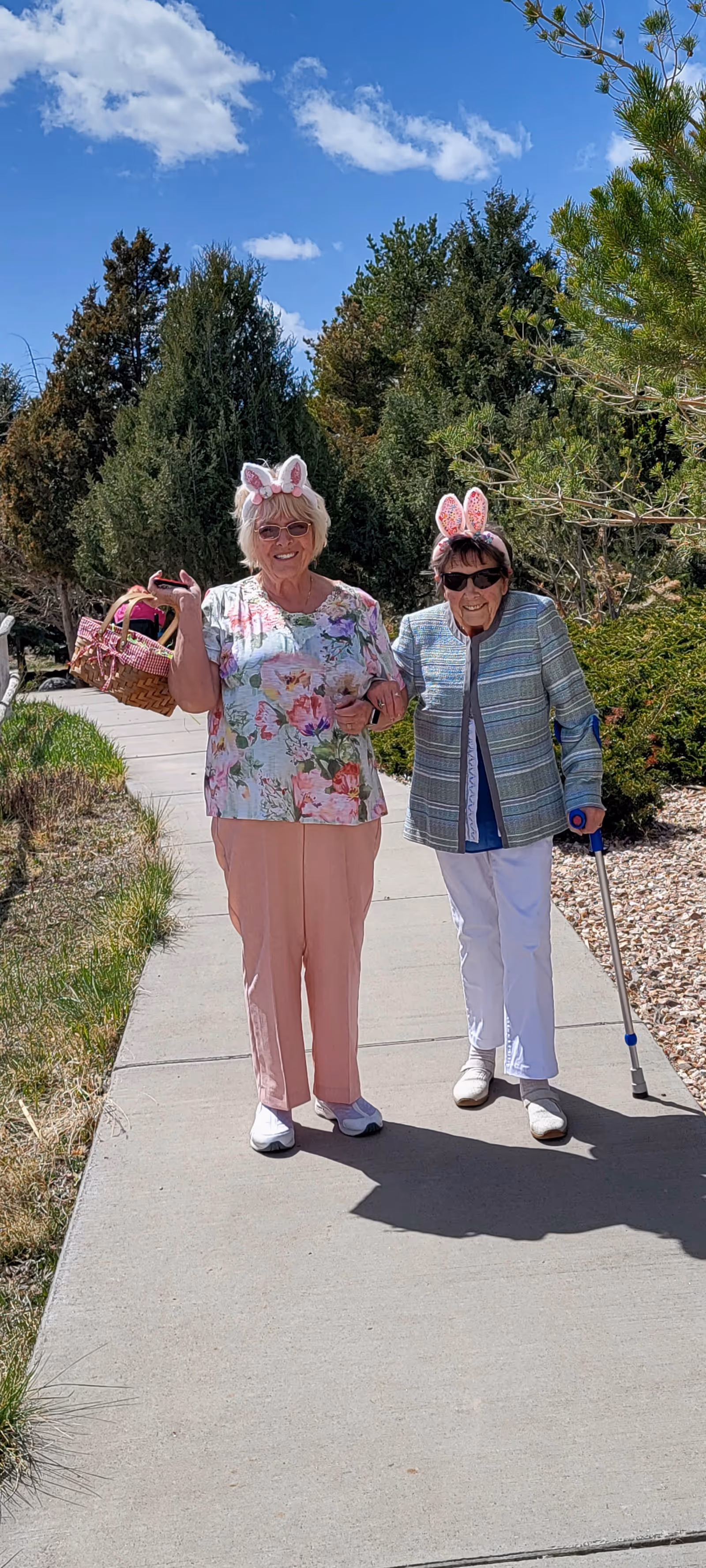 Two elderly women walking outdoors on a sunny day along a paved path surrounded by greenery. Both women are wearing bunny ear headbands and sunglasses. One woman is holding a basket and the other is using a cane.