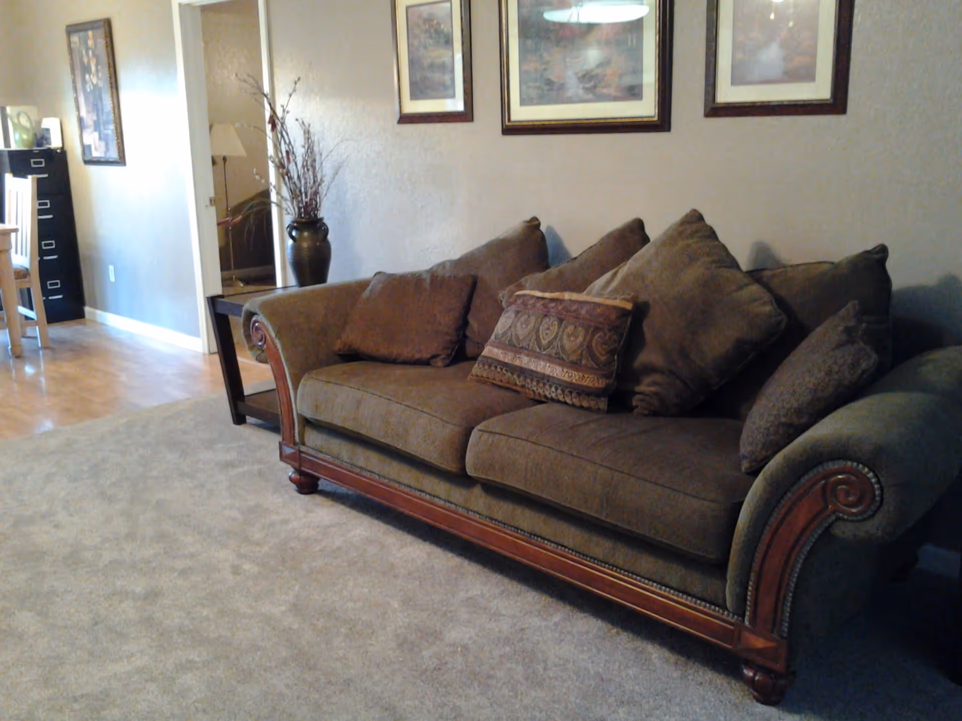 A living room area with a brown upholstered sofa adorned with multiple decorative pillows. Behind the sofa, there are three framed pictures hanging on a beige wall. To the left, there is a small table with a vase containing decorative branches, and further left, a glimpse of a dining area with a wooden chair and a black filing cabinet.