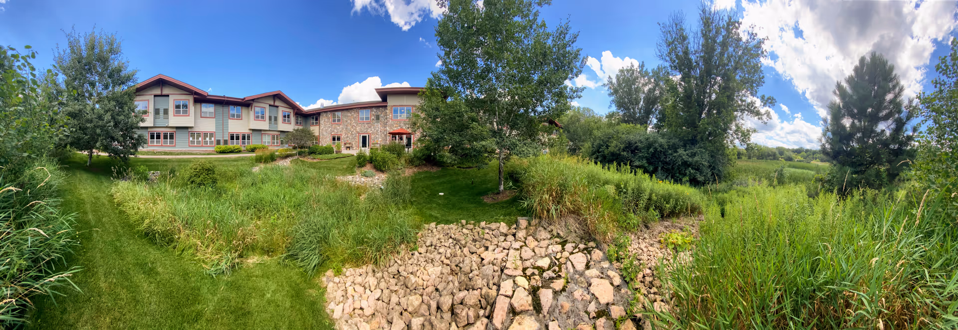 Panoramic view of the exterior of a senior living facility building with a stone and siding facade, surrounded by green grass, trees, and a rocky drainage area under a partly cloudy blue sky.