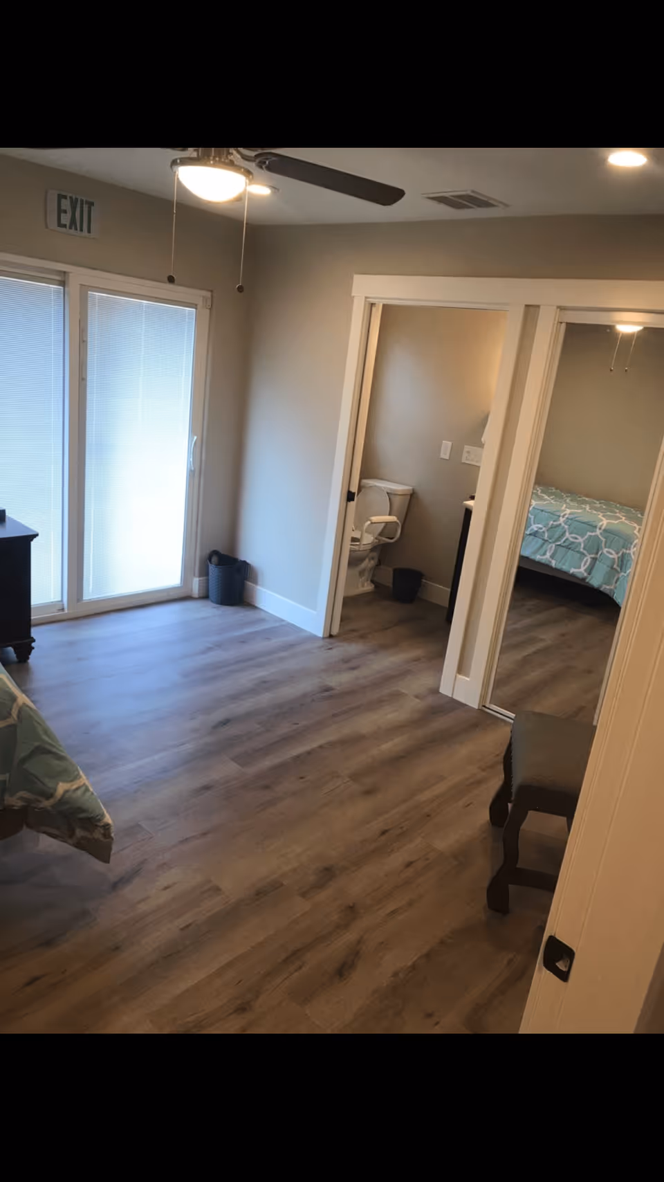Interior view of a senior living facility room with wooden flooring, a ceiling fan with light, a sliding glass door with blinds, a small bathroom with a toilet and grab bars, and a bedroom with a bed covered in a blue patterned bedspread. A small chair is visible near the entrance.
