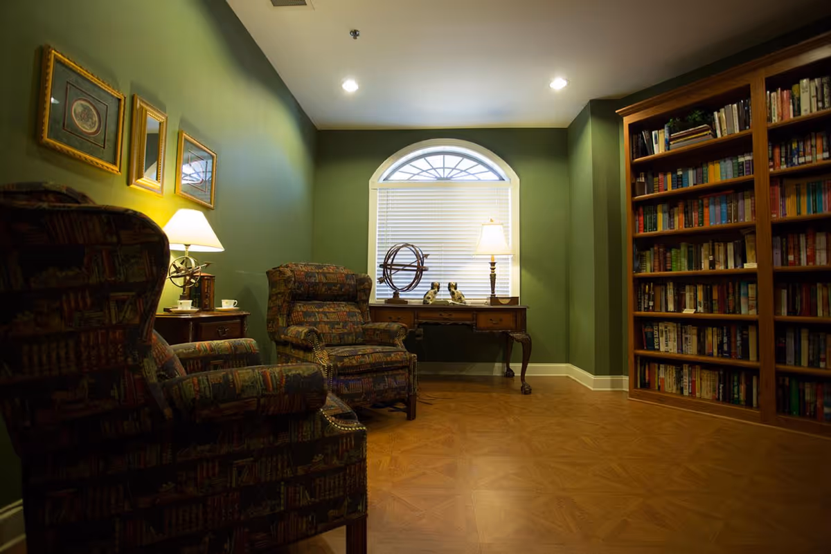 Cozy reading room with two patterned armchairs, side tables and lamps, a desk under an arched window, and a large bookshelf.