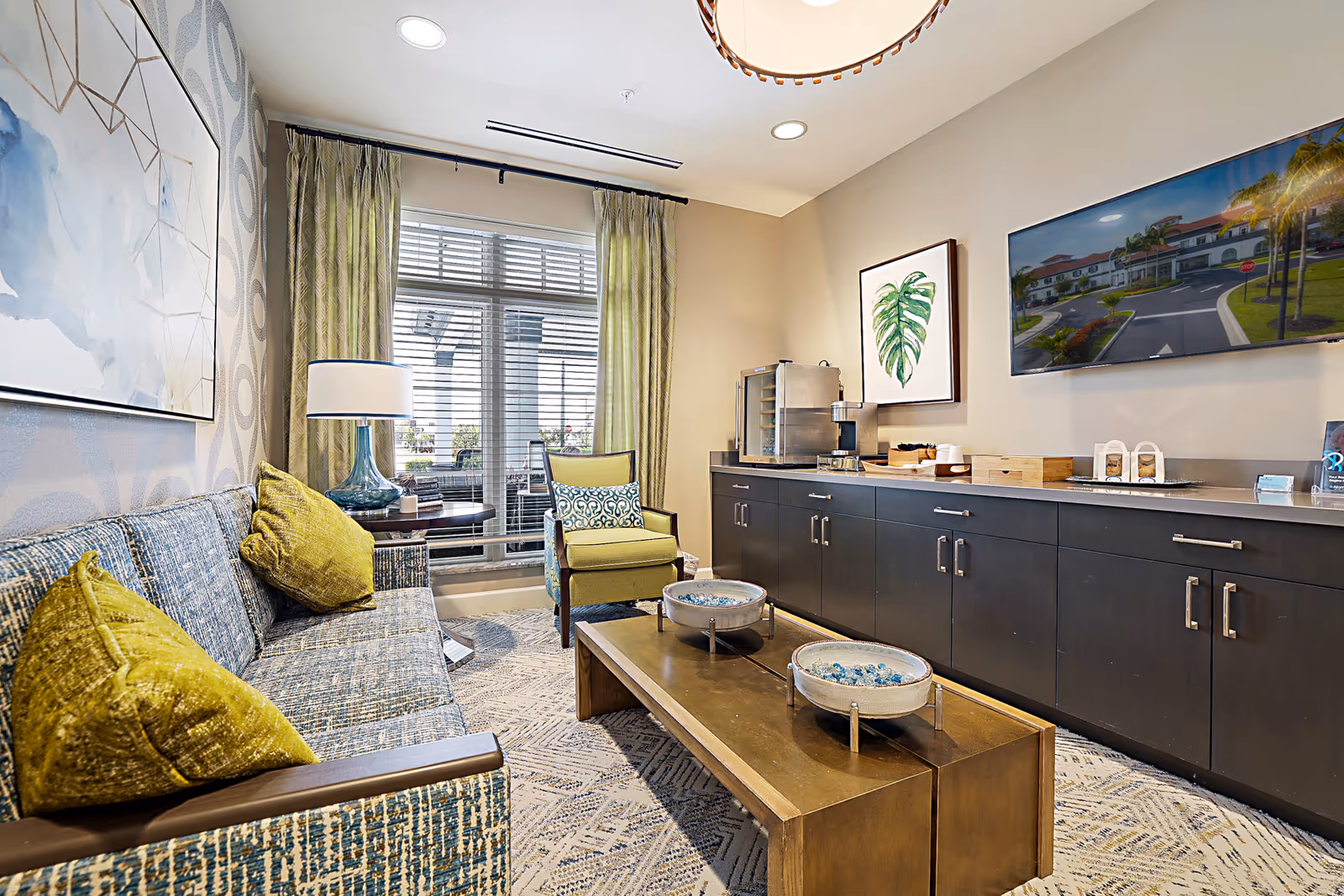 A bright senior living lounge with a patterned sofa and green pillows, an armchair, coffee table, long dark cabinets and a wall-mounted TV.