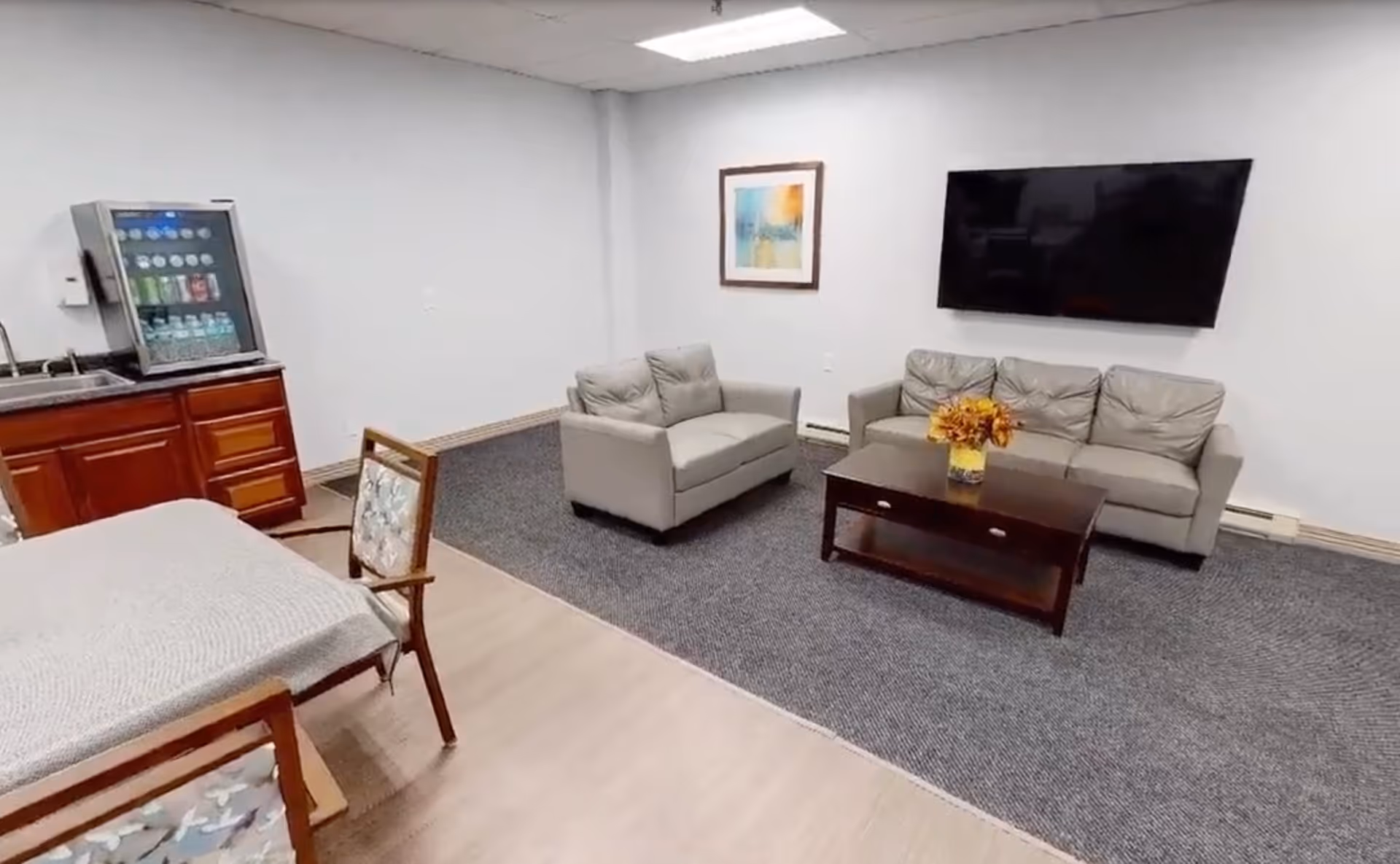 A senior living lounge with two gray sofas, a coffee table and wall-mounted TV, plus a small kitchenette/vending machine and dining chairs.