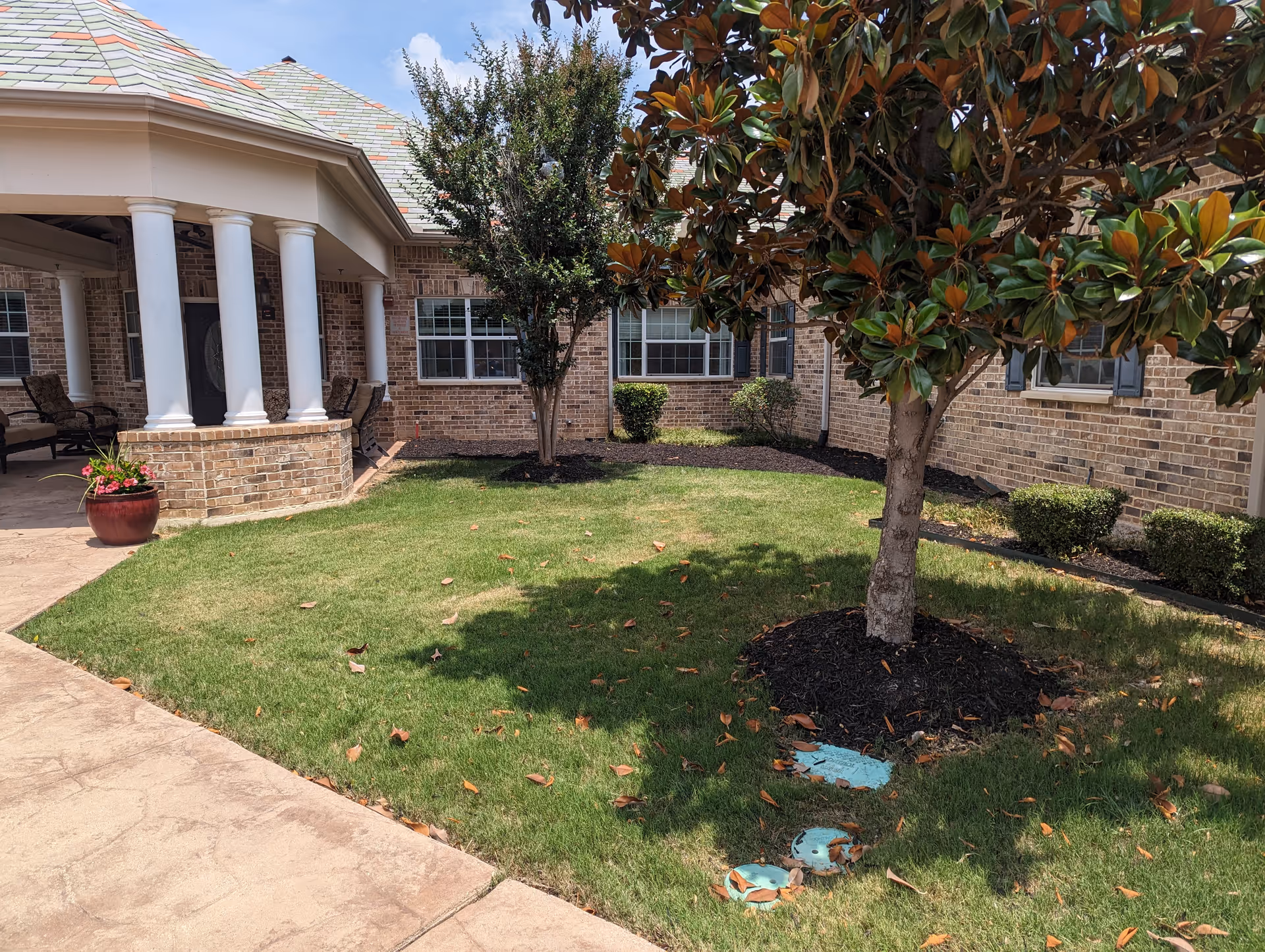 Outdoor courtyard area with green grass, a large tree, and smaller bushes in front of a brick building with white columns and a covered seating area with chairs. The sky is clear and blue.