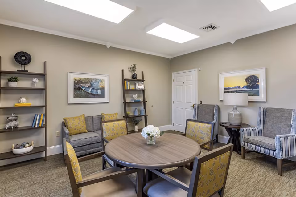 Bright common lounge with a round wooden table surrounded by upholstered chairs, armchairs by a side table, bookshelves, and framed artwork.