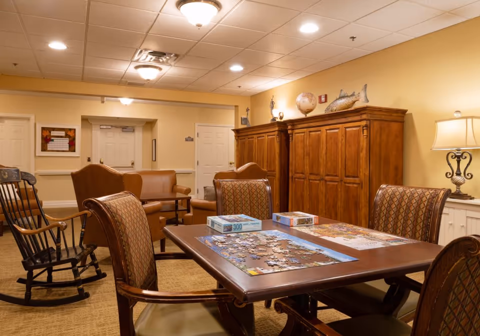A cozy common area in a senior living facility featuring a wooden table with partially completed jigsaw puzzles, surrounded by four cushioned chairs. In the background, there are two brown leather armchairs, a wooden rocking chair, and a large wooden cabinet with decorative items on top, including a globe and a fish sculpture. The walls are painted a warm beige color, and the ceiling has recessed lighting and ceiling fixtures.