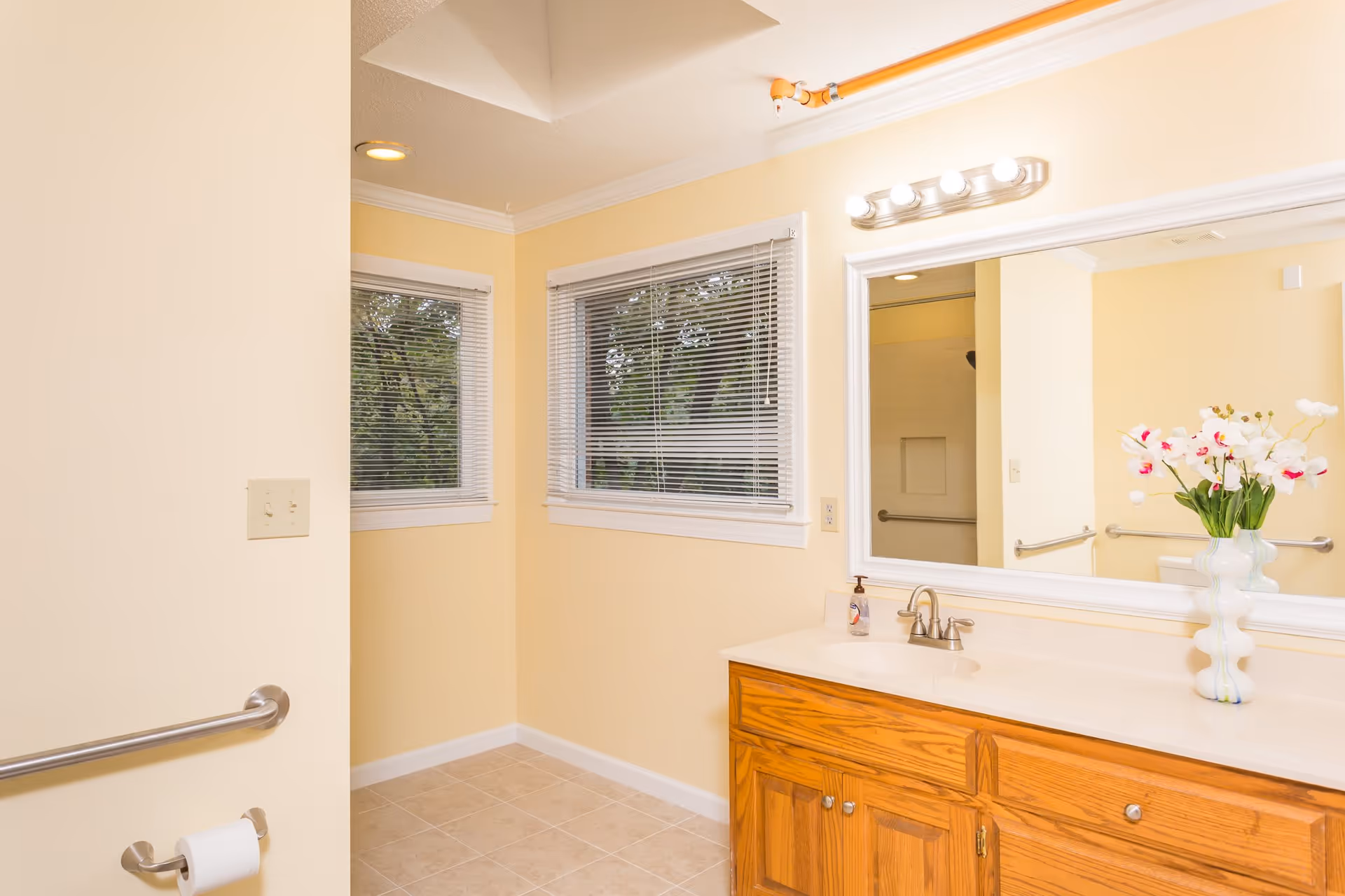 A bright bathroom with beige walls and tiled floor. There is a wooden vanity with a white countertop, a sink, and a large mirror above it. On the countertop, there is a soap dispenser and a vase with white and pink flowers. Two windows with white blinds let in natural light. A toilet paper holder with a roll of toilet paper and a grab bar are mounted on the wall.