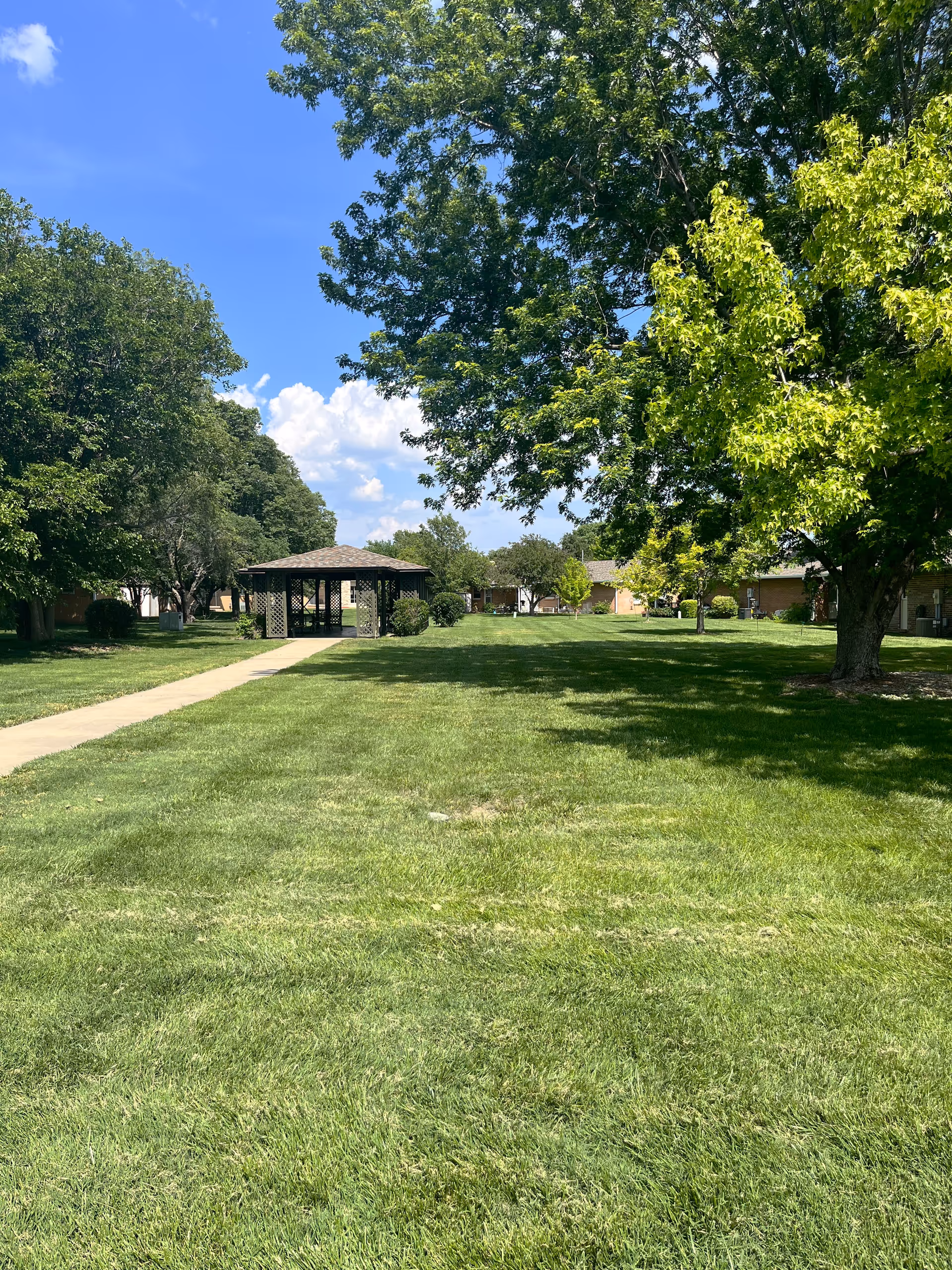 Green lawn courtyard with a central gazebo, surrounded by trees and single-story buildings under a blue sky.