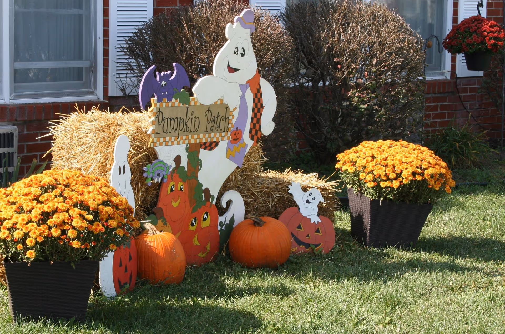 Outdoor Halloween-themed decoration featuring a hay bale, two orange pumpkins, two pots of orange flowers, and cutouts of friendly ghosts, a bat, a spider, and pumpkins with smiling faces. A sign reads 'Pumpkin Patch'. The decorations are set on a grassy lawn in front of a brick building with windows and bushes.