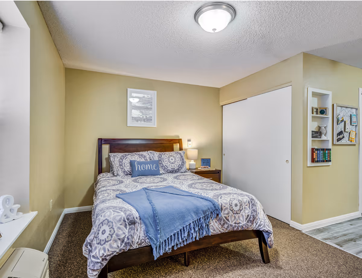 A cozy bedroom with a wooden bed frame, patterned bedding in shades of gray and white, and a blue throw blanket. There is a blue pillow with the word 'home' on it. A nightstand with a lamp and small decor items is beside the bed. The walls are painted light yellow, and there is a framed picture above the bed. A white sliding closet door is visible on the right side, along with built-in shelves holding books and decorative items.