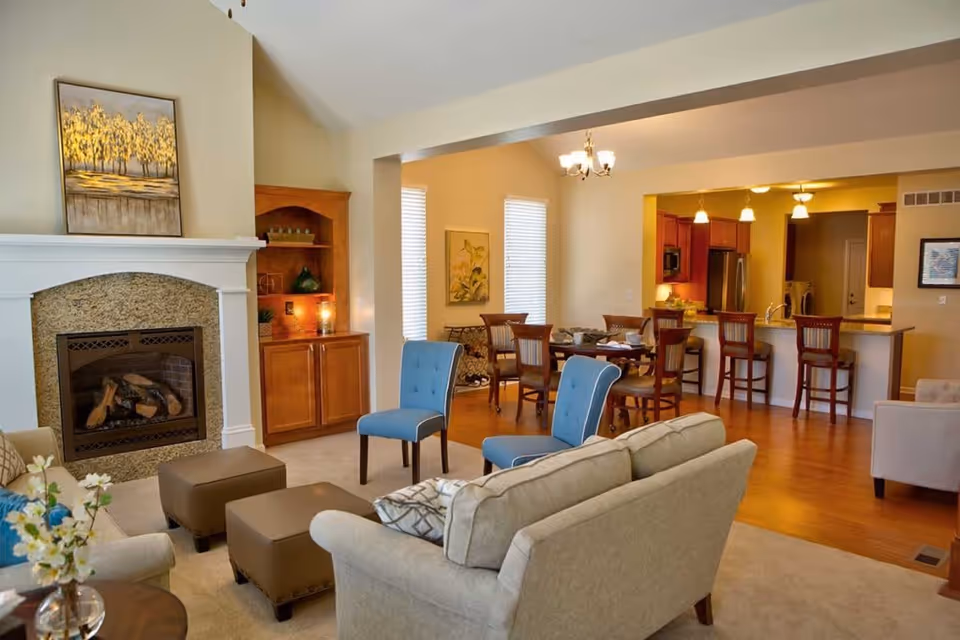 A cozy senior living community interior featuring a living room with beige sofas, two blue chairs, and two brown ottomans arranged around a fireplace with a granite surround. Behind the living room is a dining area with a wooden table and chairs, and a kitchen with a breakfast bar and stools. The space has warm lighting and wooden flooring in the dining and kitchen areas.
