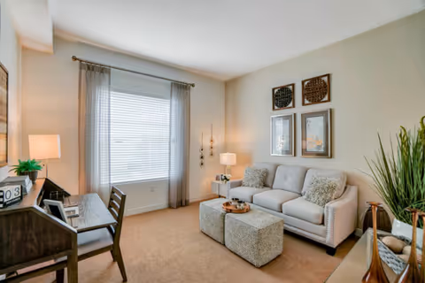 A cozy living room with a beige sofa adorned with two patterned pillows, two matching ottomans in front, a wooden desk with a chair on the left, a window with sheer curtains letting in natural light, and decorative wall art above the sofa. There are also two table lamps and a tall green plant on the right side.