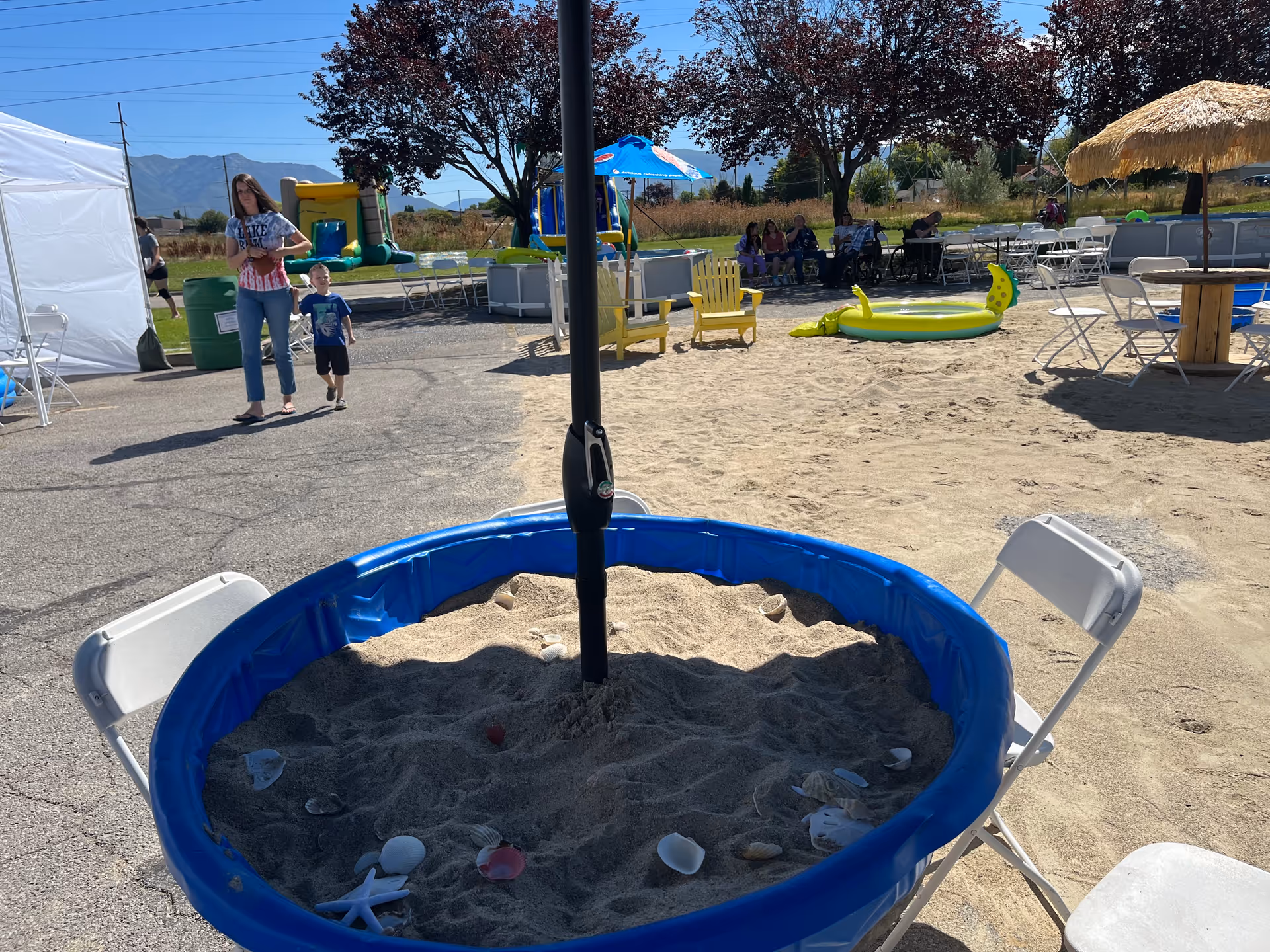 Outdoor area with a small blue plastic sandbox filled with sand and seashells, surrounded by white folding chairs. In the background, there are yellow Adirondack chairs, a small inflatable pool, white tables and chairs, a thatched umbrella, and people sitting under trees. A woman and a child are walking on a paved path. The scene is sunny with clear skies and mountains in the distance.