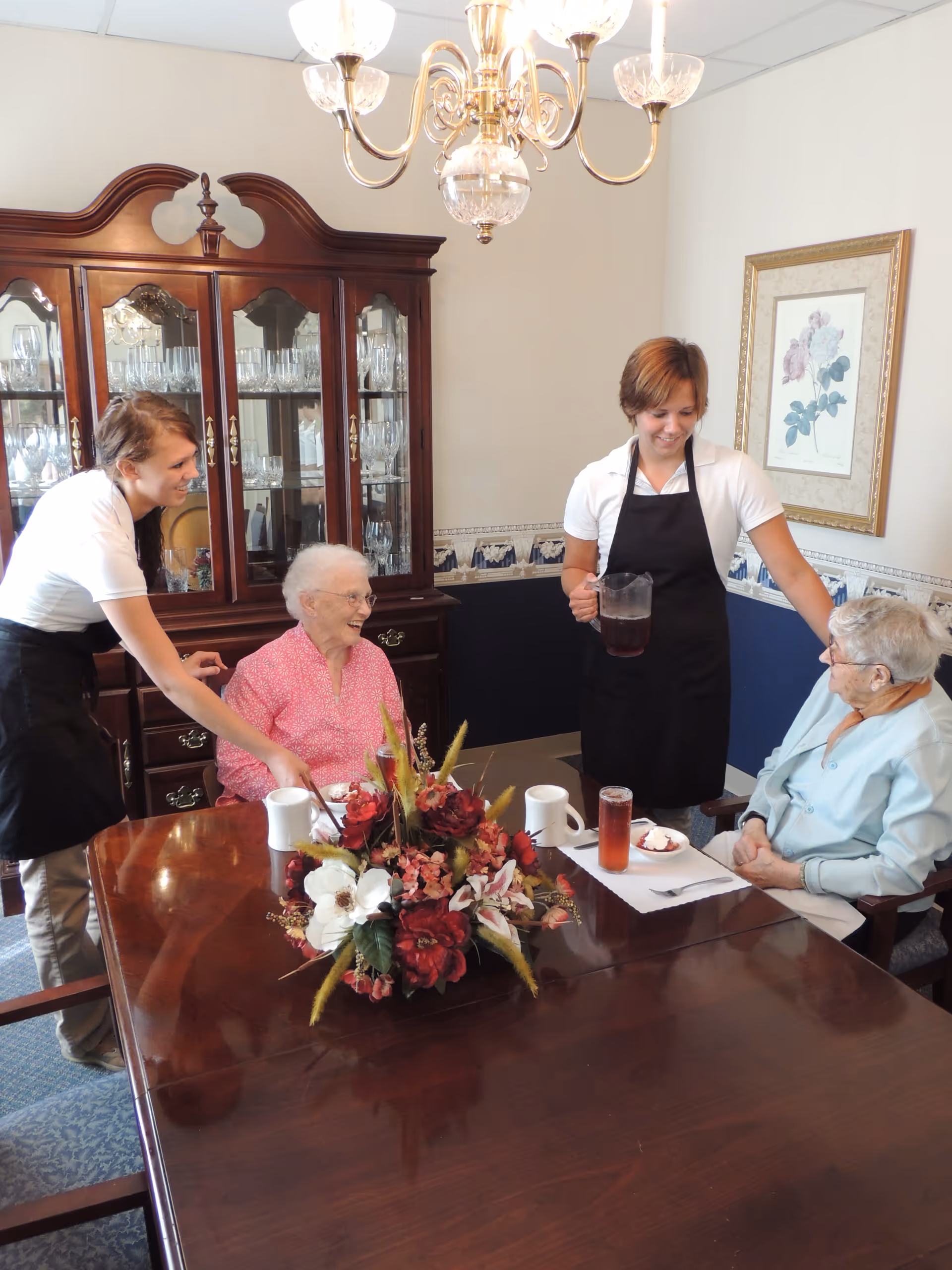 Two elderly women sitting at a wooden dining table with a floral centerpiece, being served by two caregivers wearing white shirts and black aprons in a dining room with a chandelier and a glass cabinet filled with glassware.
