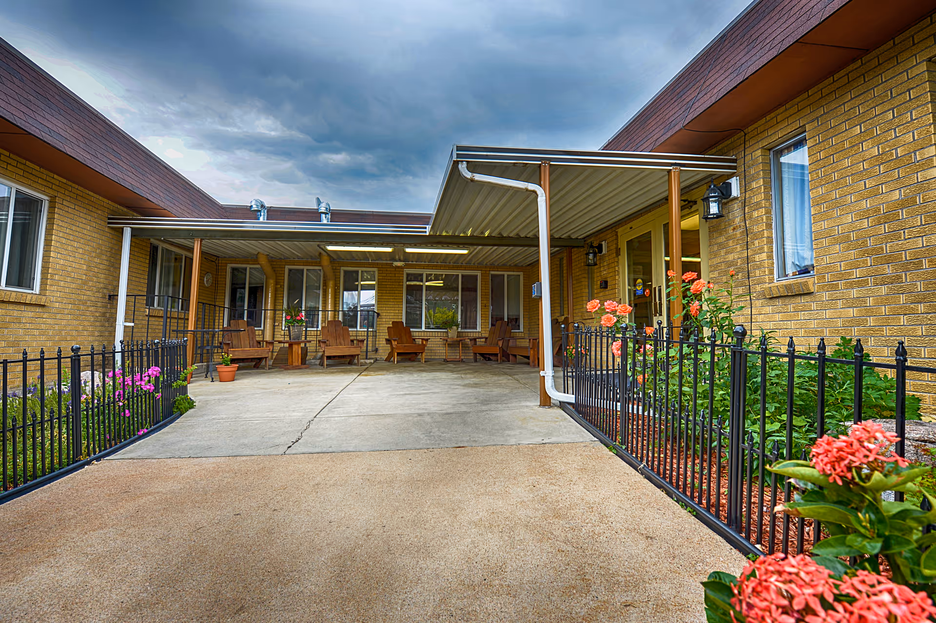 Outdoor patio area of a senior living facility with a covered seating area featuring wooden chairs and small tables. The patio is surrounded by a black metal fence with flower beds containing pink and orange flowers. The building has yellow brick walls and several windows, with a cloudy sky overhead.