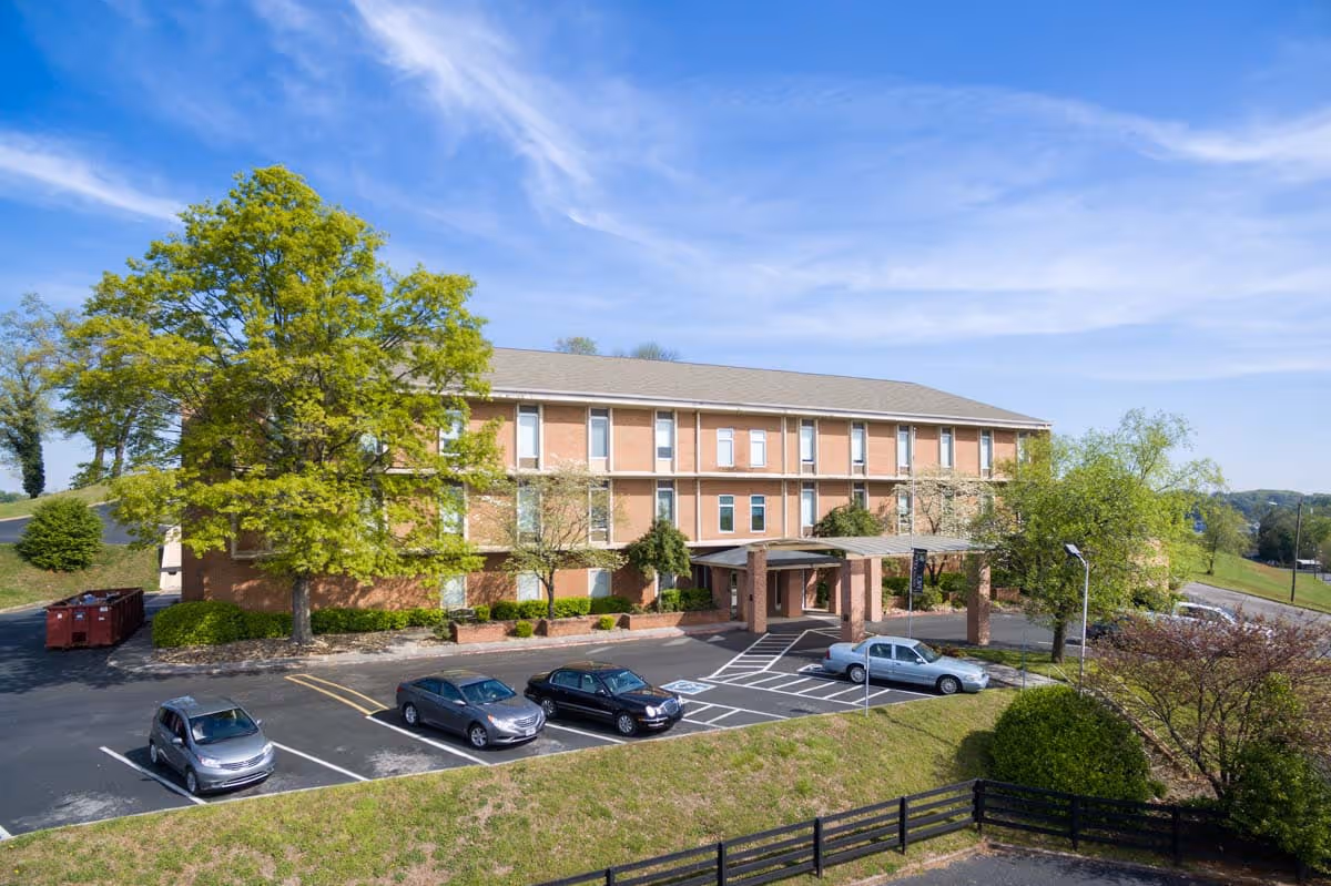 Exterior view of a three-story brick building with a covered entrance, surrounded by trees and a parking lot with several cars parked. The sky is clear with some wispy clouds.