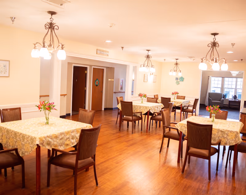 A bright dining room with wooden floors, several tables covered with patterned tablecloths, each adorned with a small vase of flowers. The room has multiple hanging light fixtures and wooden chairs around the tables. Doors and a seating area with armchairs and windows are visible in the background.