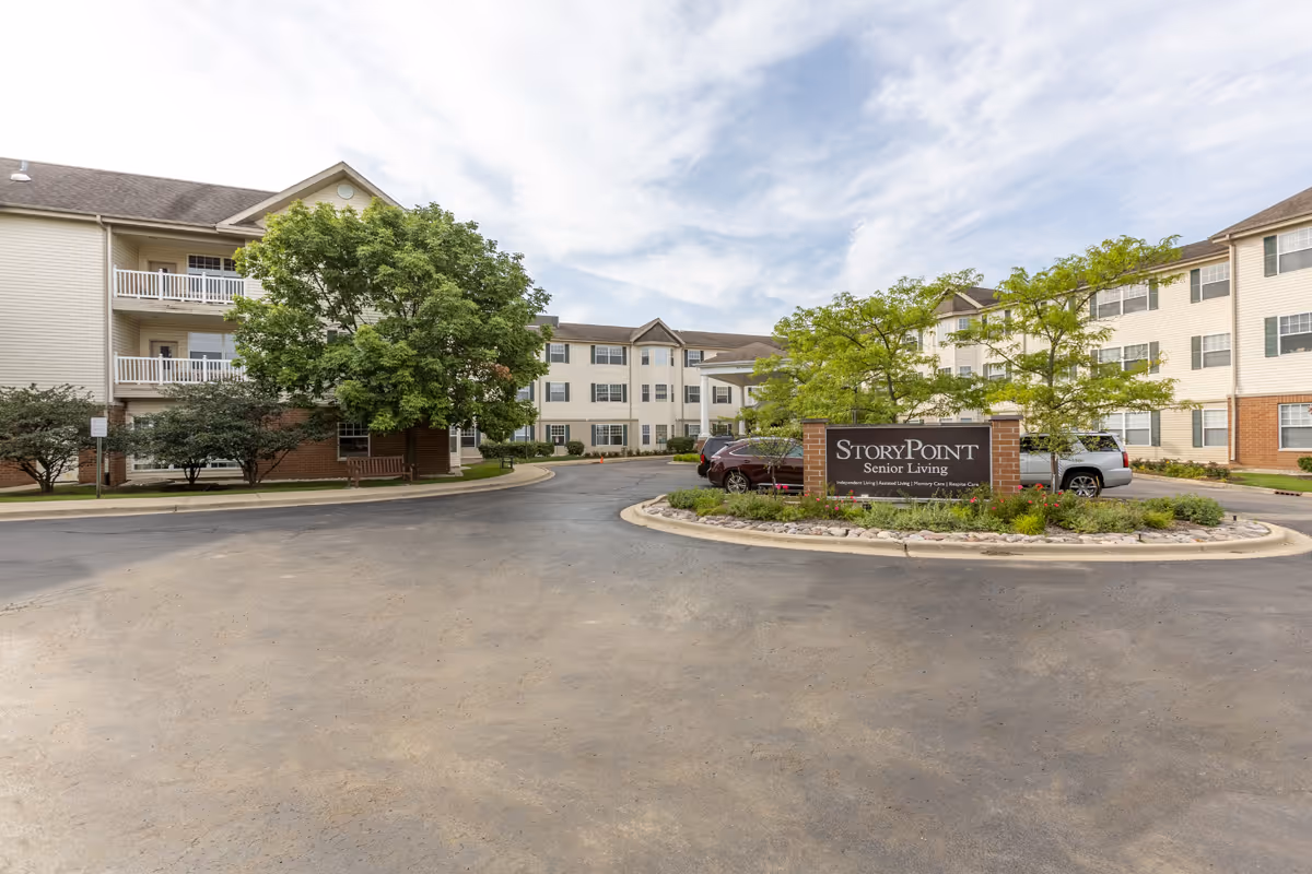 Exterior view of StoryPoint Libertyville senior living facility showing a three-story building with balconies, trees, parked cars, and a sign reading 'StoryPoint Senior Living' at the entrance.