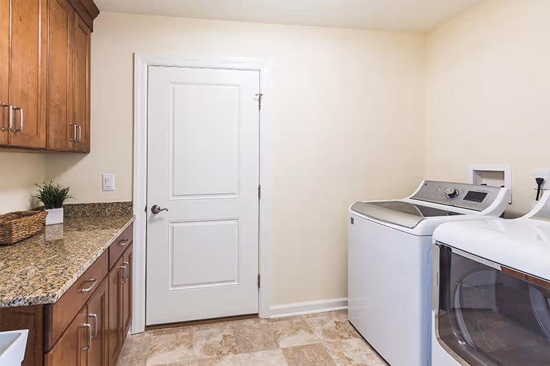 Laundry room with a washer and dryer on the right, wooden cabinets and a granite countertop on the left, and a closed white door centered.