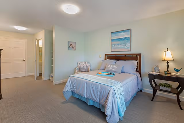 A neatly made bed with light blue bedding in a senior living facility bedroom. There is a wooden headboard, a side table with a lamp, clock, and decorative items, and a cushioned chair with a pillow that says 'Home Sweet Home'. A painting of a beach scene hangs above the bed. The room has light-colored walls and carpeted floor, with a door leading to another room or bathroom.