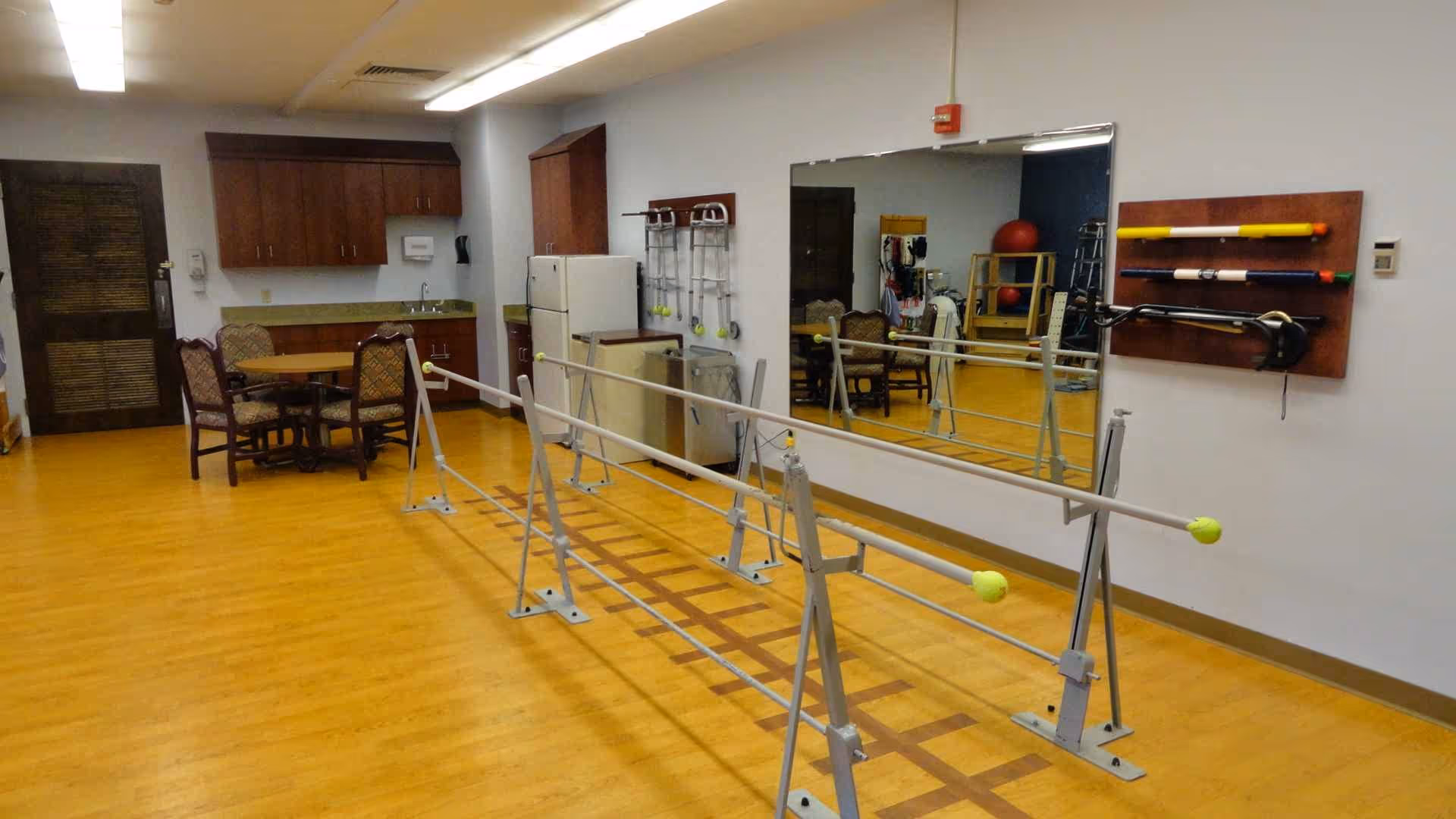 A room with wooden flooring featuring parallel bars used for physical therapy or rehabilitation. There is a large mirror on one wall, a small kitchenette with cabinets, a refrigerator, and a table with four chairs. Various exercise equipment is mounted on the wall next to the mirror.