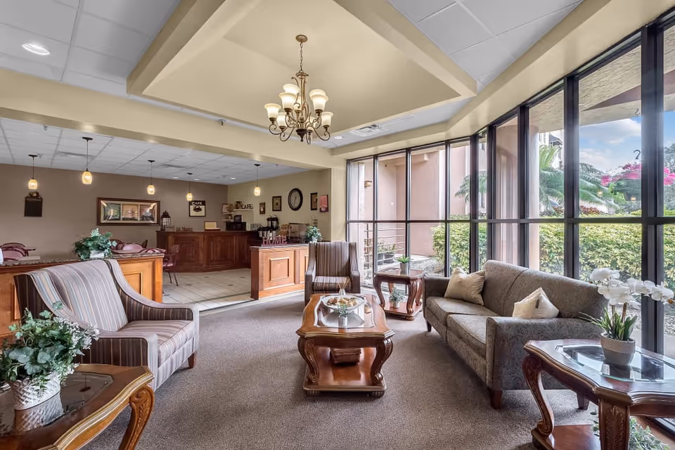 Bright sitting area with sofas, armchairs, wooden coffee tables and large floor-to-ceiling windows overlooking greenery.