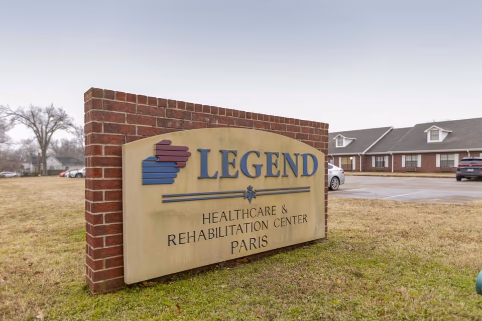 A large stone and brick sign outside a building that reads 'LEGEND Healthcare & Rehabilitation Center Paris' with a parking lot and part of the facility building visible in the background.