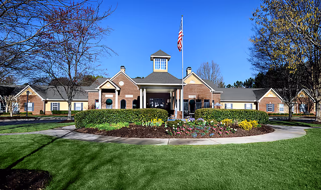 Front exterior view of Belmont Village Senior Living Johns Creek building with a circular driveway, landscaped garden with flowers and bushes, an American flag on a flagpole, and clear blue sky.
