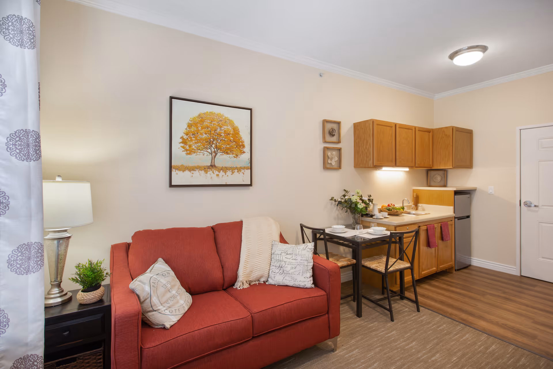 A cozy living area featuring a red loveseat with two decorative pillows and a cream throw blanket. To the left is a side table with a lamp and a small green plant. Above the loveseat hangs a framed picture of a tree with orange leaves. To the right is a small dining table set for two with plates and cups, adjacent to a kitchenette with wooden cabinets, a sink, and a mini refrigerator. The room has beige walls and wood flooring near the kitchenette, with carpet in the living area.