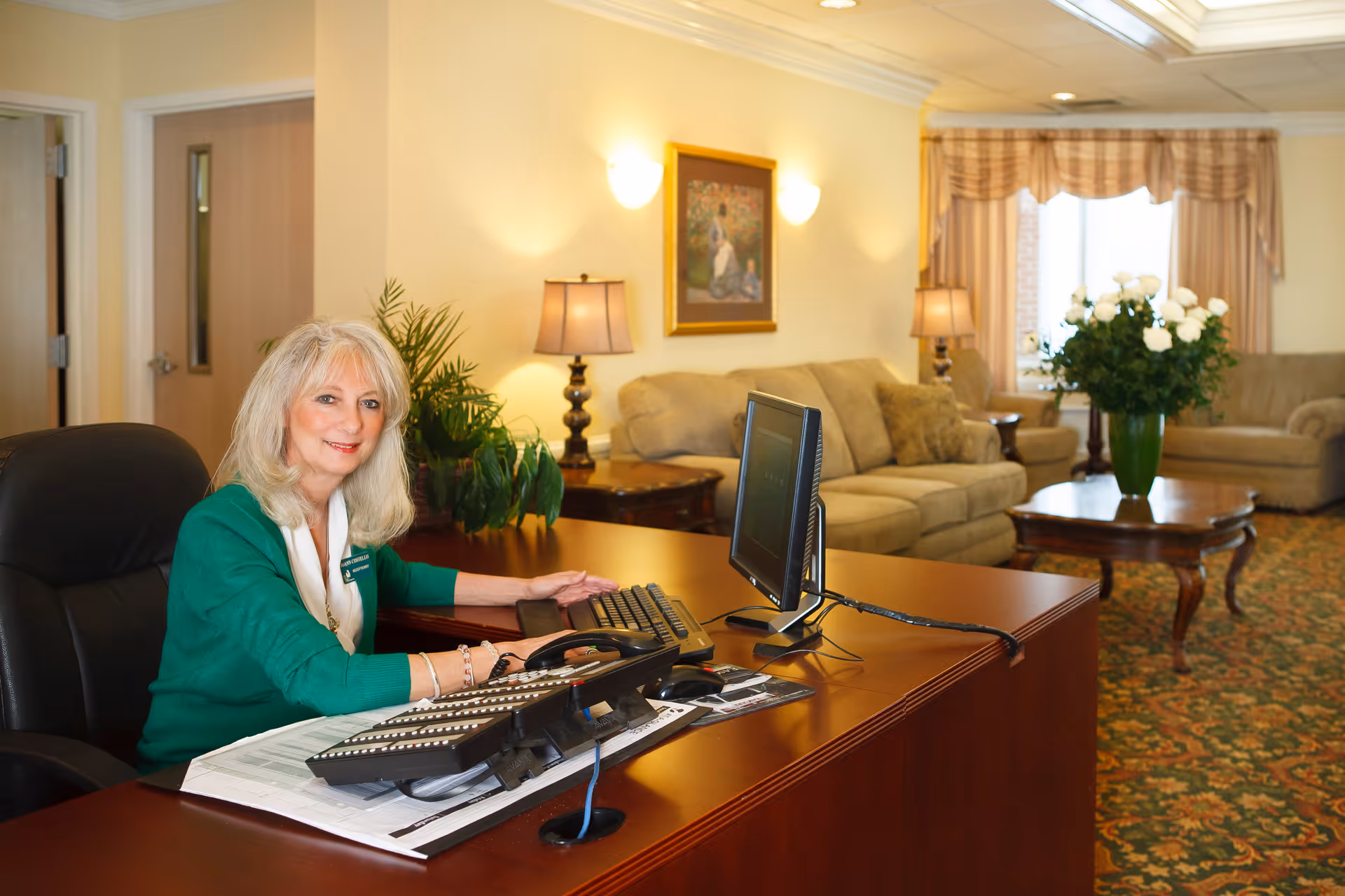 A woman with blonde hair wearing a green cardigan sits at a reception desk with a computer and telephone in a warmly lit room. Behind her is a comfortable living area with beige sofas, a wooden coffee table with a vase of white flowers, table lamps, a framed painting on the wall, and a window with curtains.