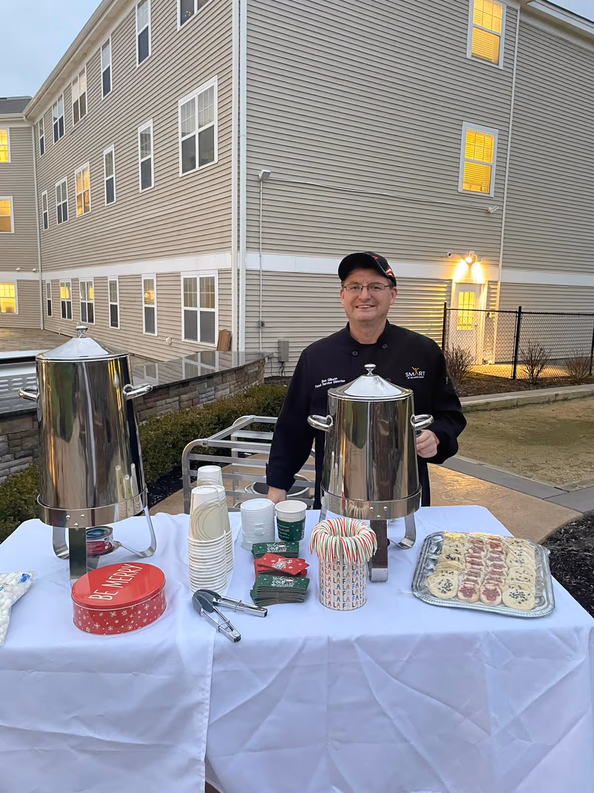 A man wearing a black chef's jacket and cap stands behind a table set up outdoors with two large silver beverage dispensers, stacks of disposable cups, holiday-themed napkins, a container holding candy canes, and a tray of assorted cookies. The background shows a multi-story building with lit windows and a fenced area.