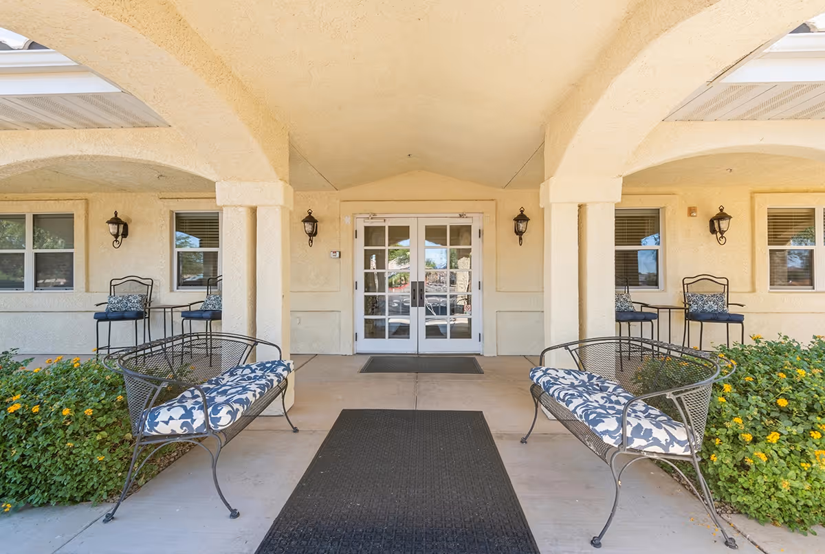 Entrance area of Trustwell Living at Kingswood Place featuring a covered walkway with two cushioned metal benches on either side, two sets of metal chairs with small tables near windows, beige stucco walls, and double glass doors in the center.
