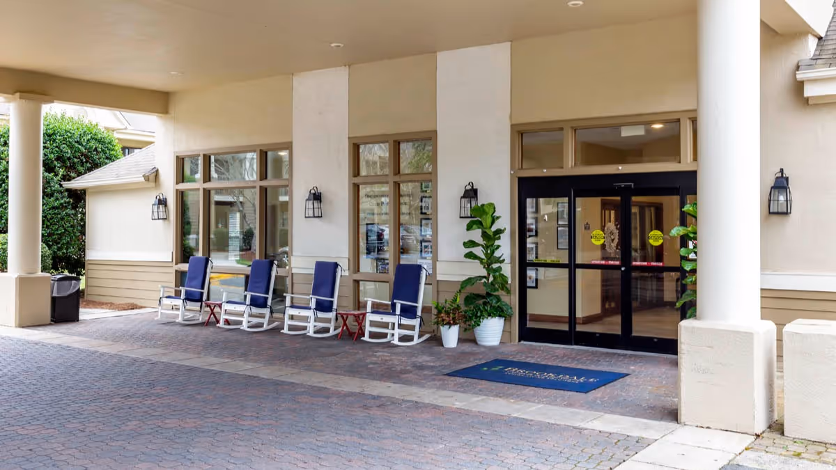 Entrance area of a senior living facility with a covered walkway, four blue rocking chairs with white frames, small red side tables, potted plants, and glass double doors leading inside.