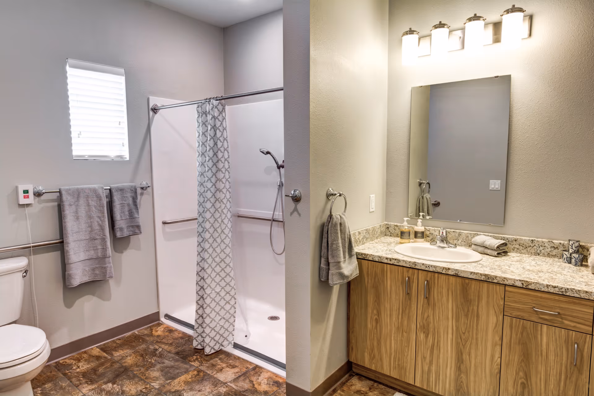 A clean and modern bathroom featuring a walk-in shower with a patterned curtain, a toilet, and a vanity with a granite countertop, sink, and mirror. The bathroom has light-colored walls, a small window with blinds, and several gray towels hanging on racks.