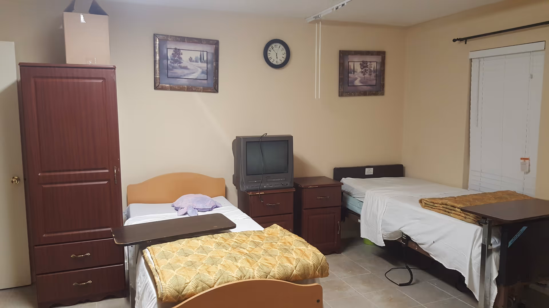 A furnished assisted-living bedroom with two single beds, a TV on a dresser, wardrobe, overbed tables, a wall clock and framed pictures.