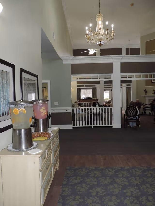 Interior view of a senior living facility common area with a chandelier hanging from the ceiling. In the foreground, there is a sideboard with two beverage dispensers containing lemon and pink drinks, along with a plate of pastries and bananas. In the background, several elderly people are seated and conversing in a lounge area with large windows and a fireplace.