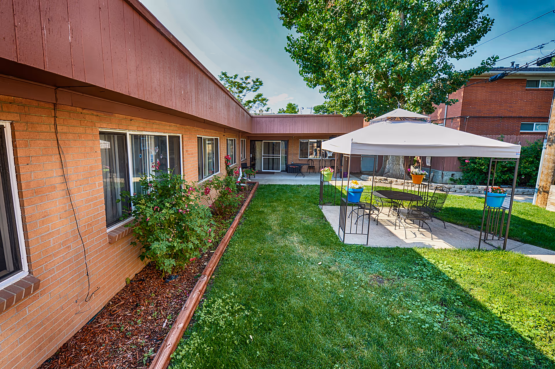 A grassy courtyard with a brick single-story building, a shaded patio gazebo with table and chairs, and potted plants.