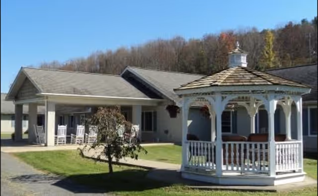 White gazebo on a lawn in front of the covered entrance to a single-story senior living building with rocking chairs on the porch.