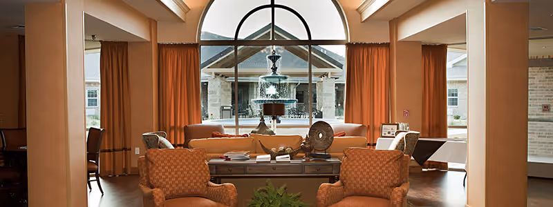A cozy living room area with two patterned armchairs and a sofa arranged around a wooden coffee table. Behind the seating area is a large arched window with orange curtains, showcasing an outdoor fountain and part of the building exterior.