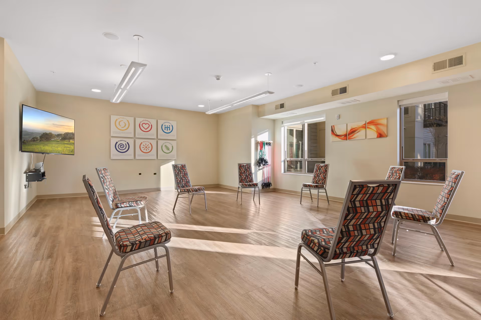 A bright, spacious room with wooden flooring and beige walls, arranged with chairs spaced evenly in a circle. The room has large windows letting in natural light, a wall-mounted TV displaying a scenic landscape, and colorful abstract artwork on the walls. Exercise bands hang near one window, suggesting the room is used for group activities or fitness classes.